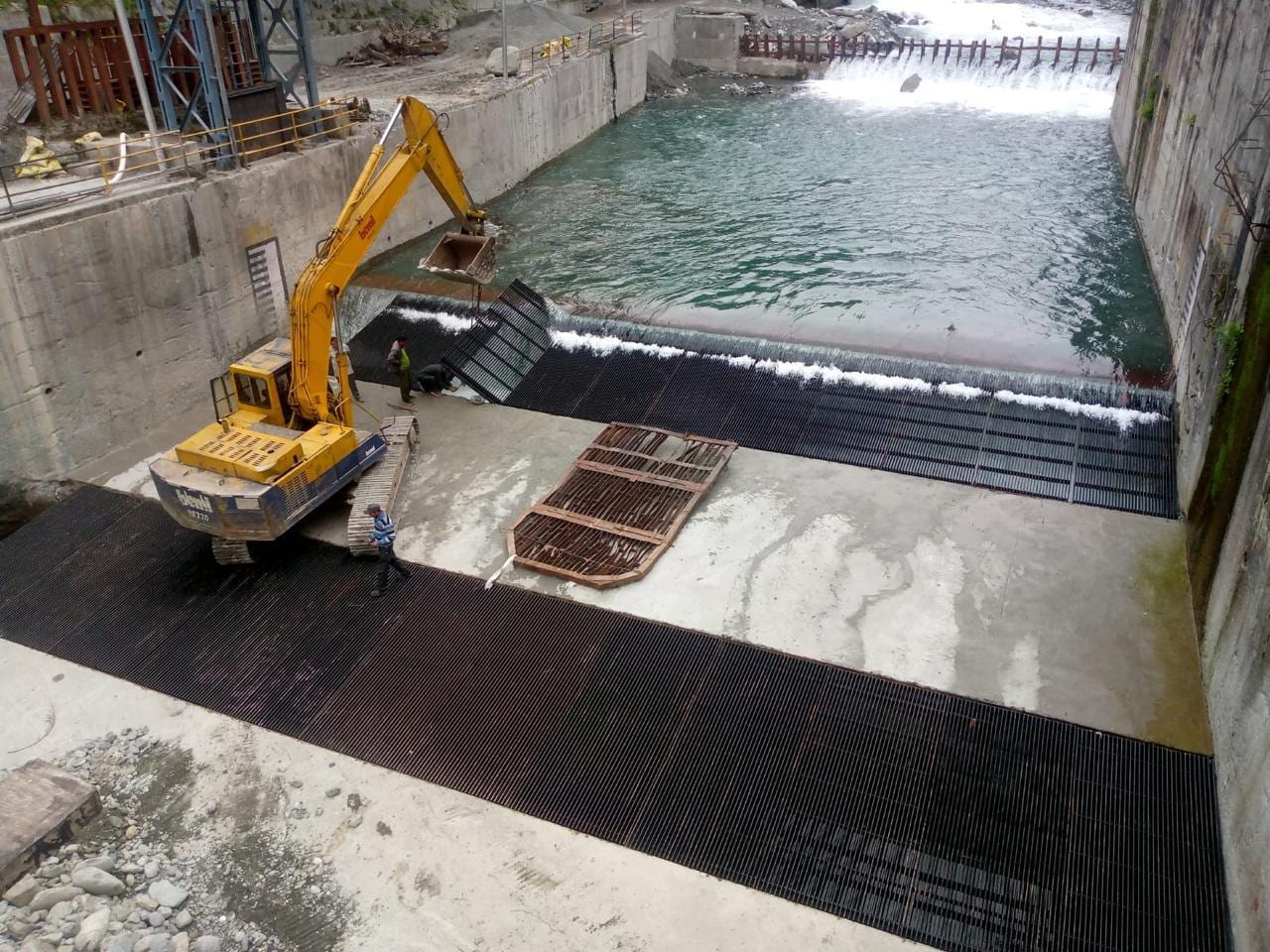 Excavator placing black trash rack panels on the concrete apron of a river weir, with water flowing over the weir behind