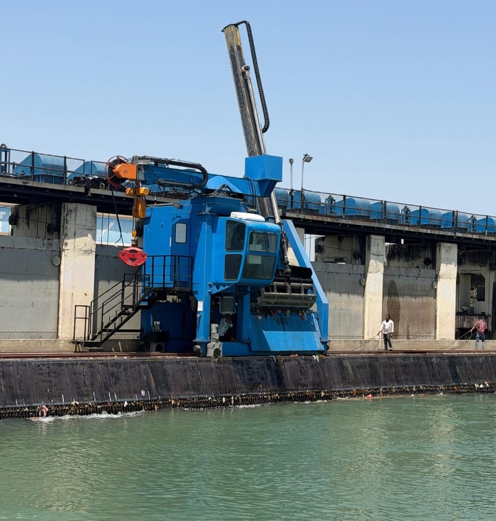 Blue trash rack cleaning machine on rails at a dam intake, with the grab raised
